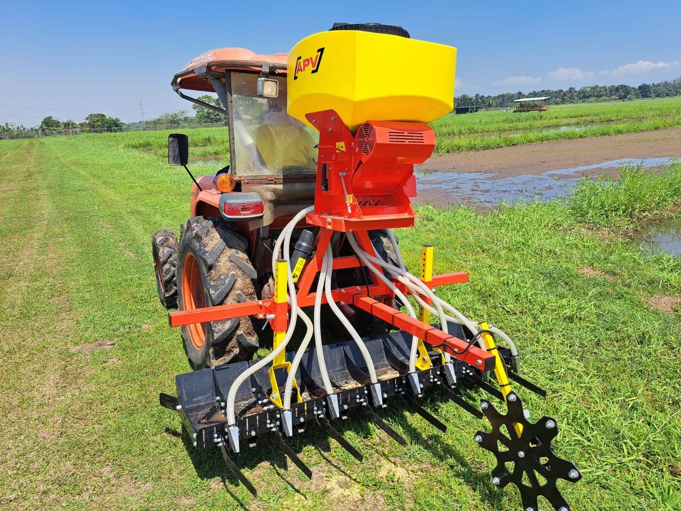 Tractor compacto rojo con tolva APV y barra sembradora sobre pastizal verde junto a un canal de riego