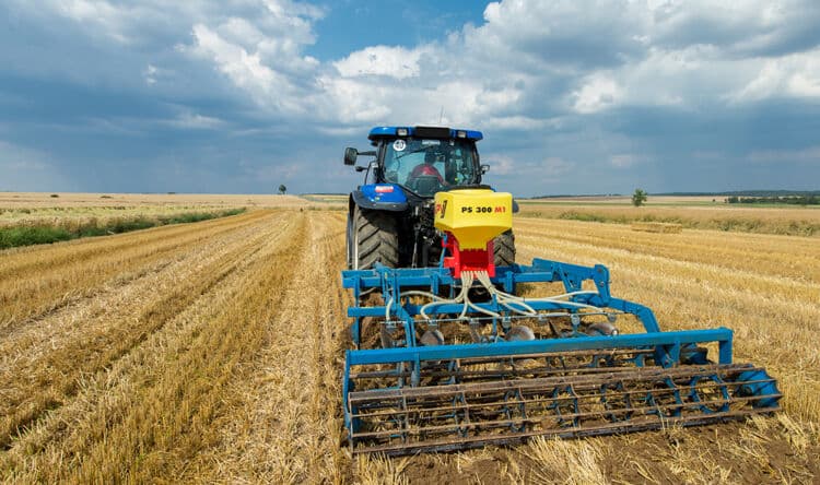 Sembradora APV PS 300 M1 sobre rastra con rodillo, tirada por tractor azul en un rastrojo de cereal recién cosechado
