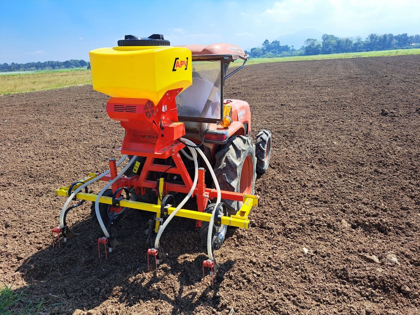 Tractor compacto rojo con barra sembradora y tolva APV sobre suelo recién preparado, montañas al fondo