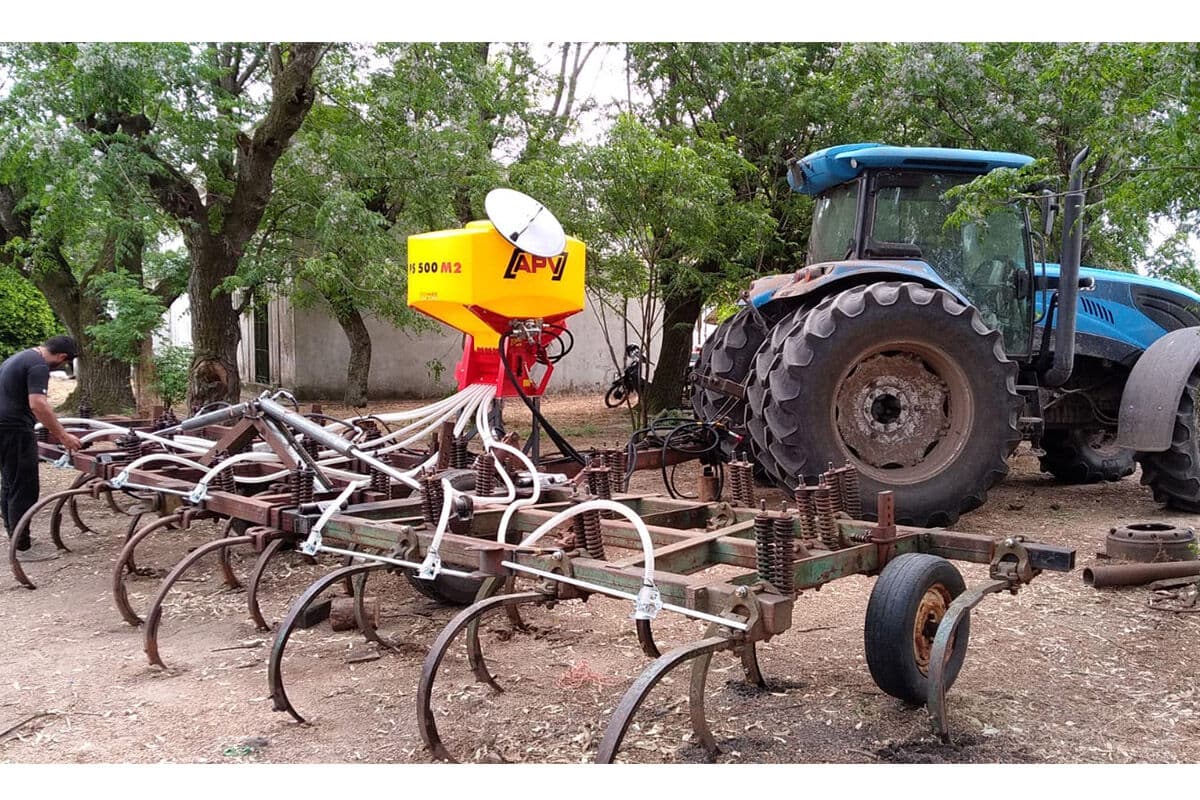 Tolva APV sobre un arado de cinceles enganchado a un tractor azul, bajo los árboles