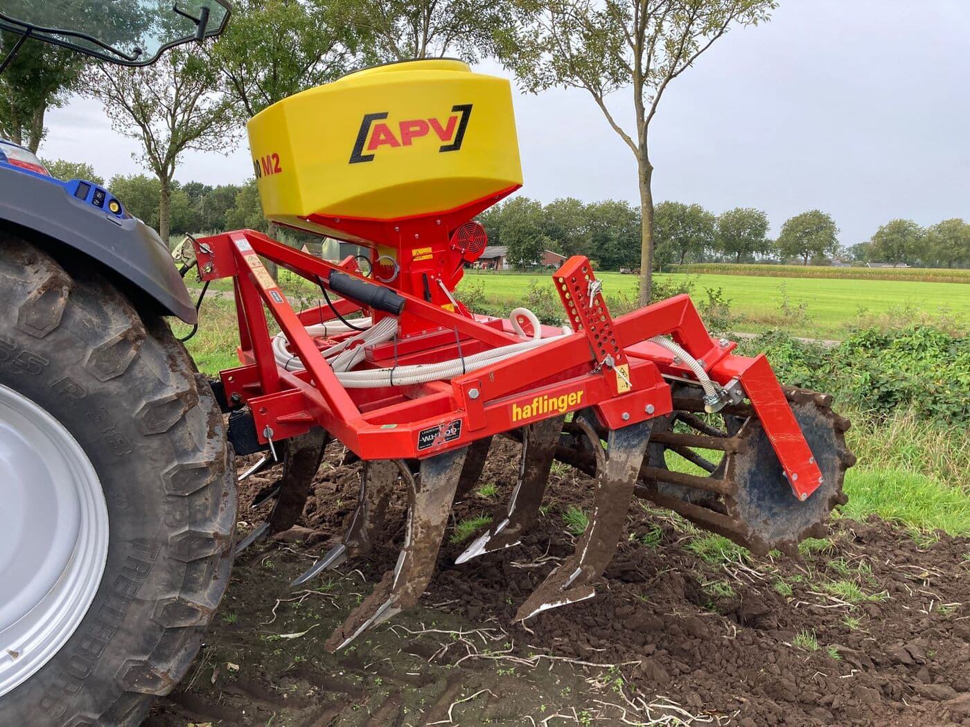 Tolva APV sobre un cultivador de brazos rígidos con tractor rojo, en campo